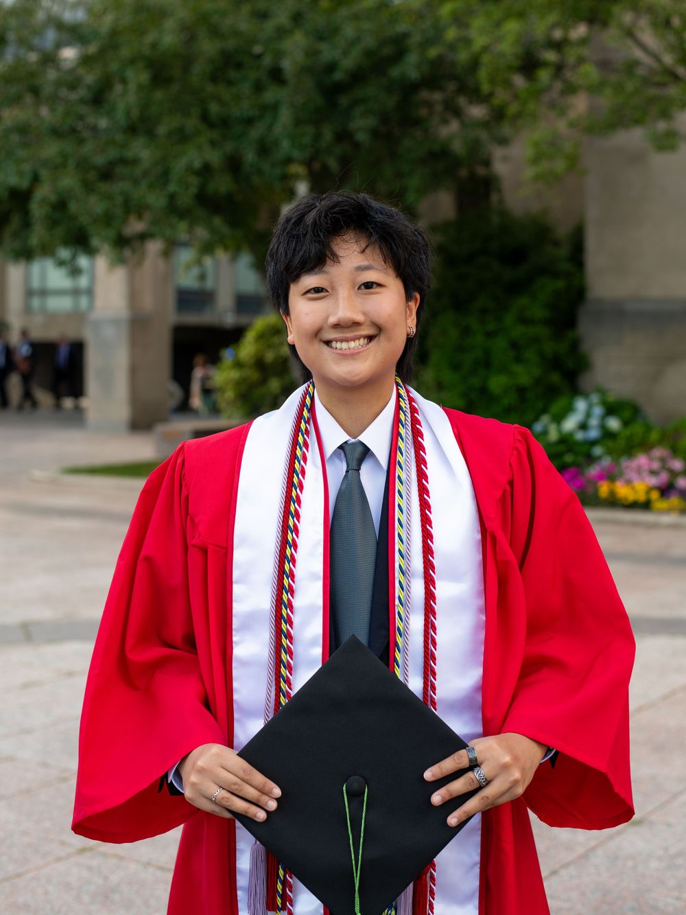 Smiling graduate in red gown holding mortarboard cap with honor cords outdoors at graduation ceremony.