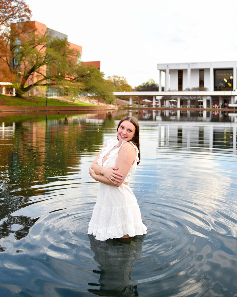 Someone wearing white dress stands in lake water with brick buildings and trees in background.