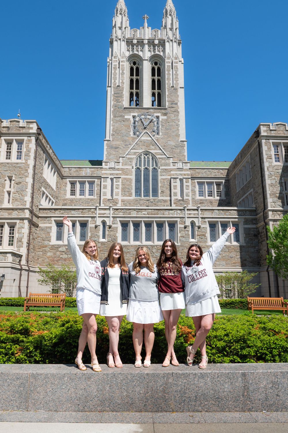 Students in white dresses celebrate graduation in front of Boston College's Gothic architecture on a sunny day.