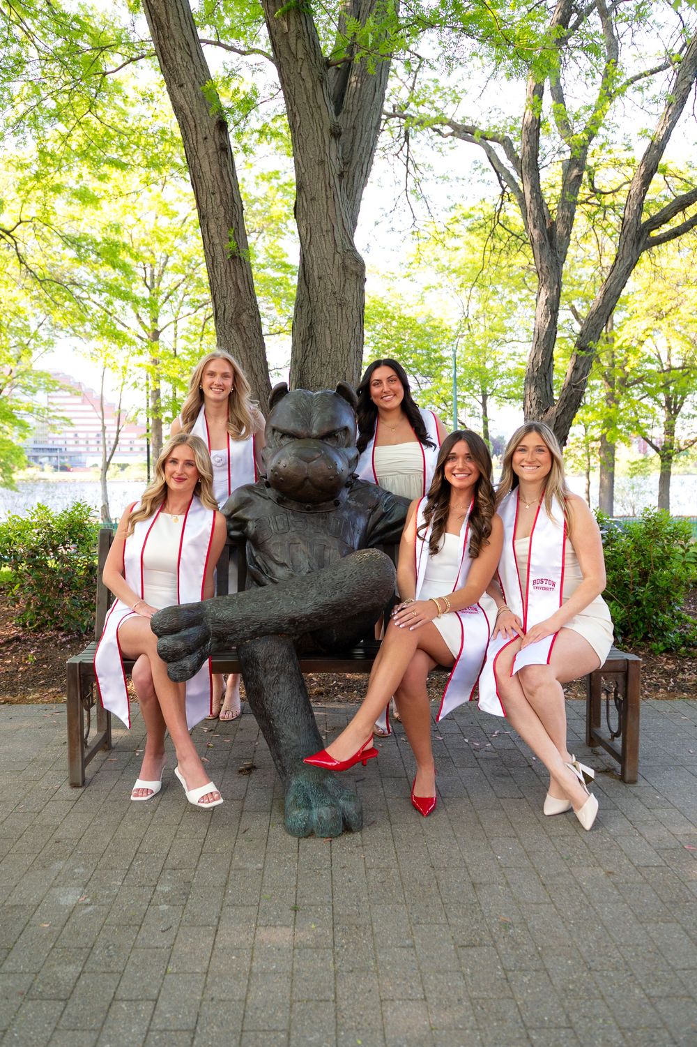 Five people sit on a bench with a bronze sculpture of a character in casual dress on a brick path with trees in the background.