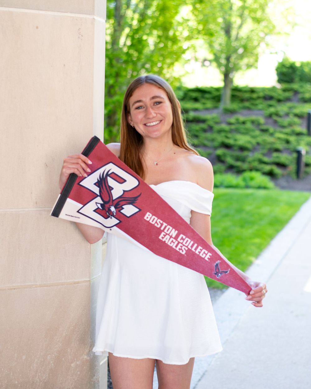 Graduate in white off-shoulder dress holds burgundy college pennant while posing next to stone column outdoors.