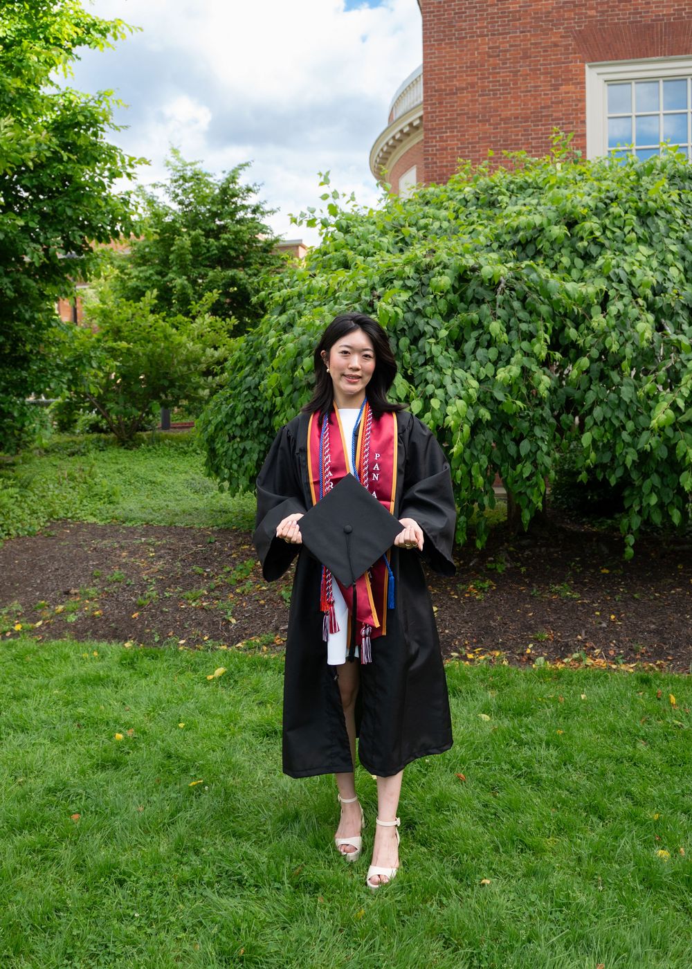Graduate in black cap and gown with colorful stoles poses on green campus lawn with trees and brick building.