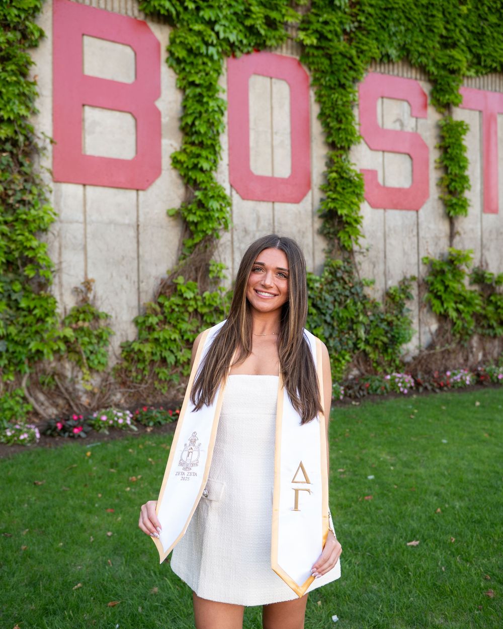 A graduate in a white dress poses by a wall with pink BOST lettering and green ivy on a sunny day.