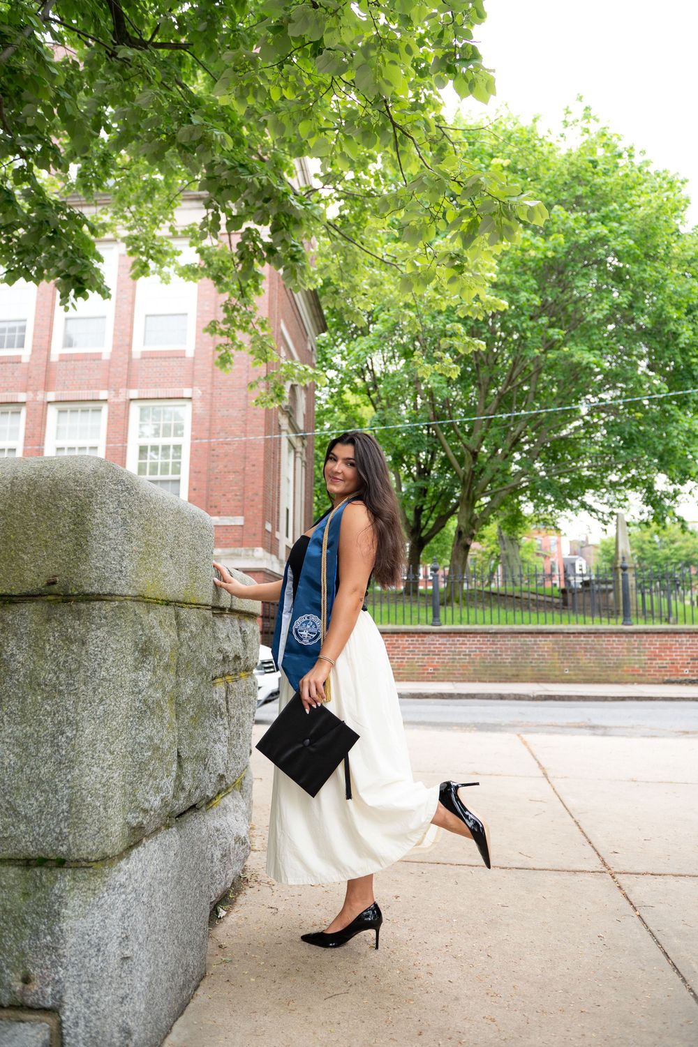 Woman in elegant white dress and denim vest posing against stone wall on city sidewalk with lush green trees.