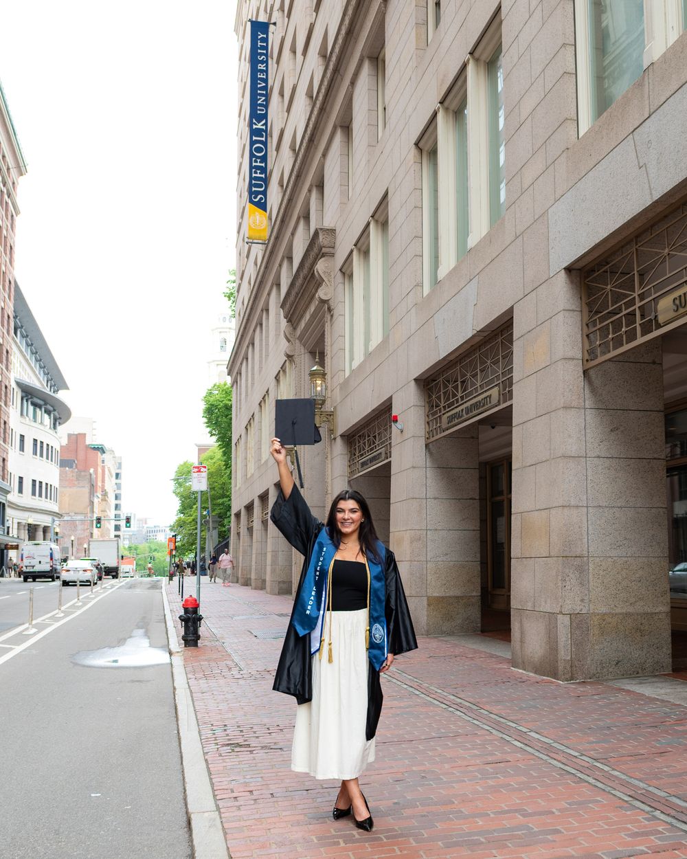 Recent graduate celebrates in cap and gown while posing on city street near Suffolk University building in downtown Boston.