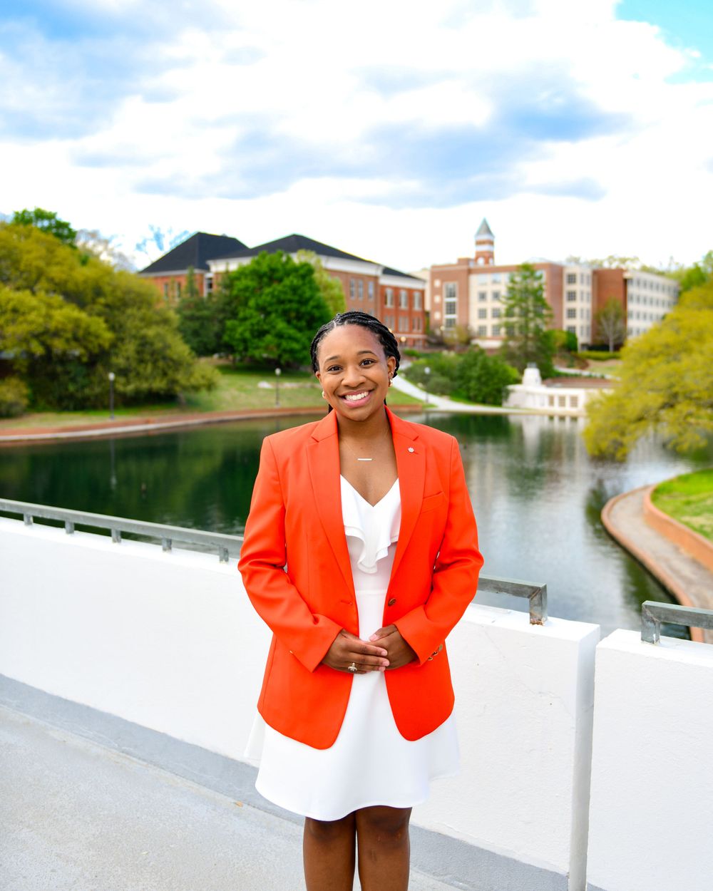 Person in orange blazer and white dress smiles by campus lake with buildings in background.