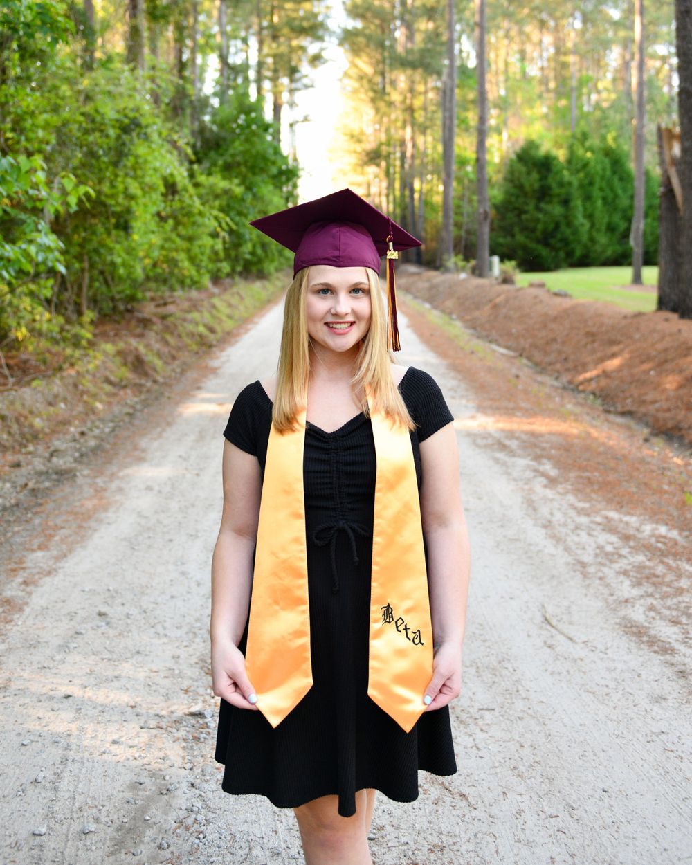 Graduate in black dress and purple cap with gold sash poses on tree-lined path.