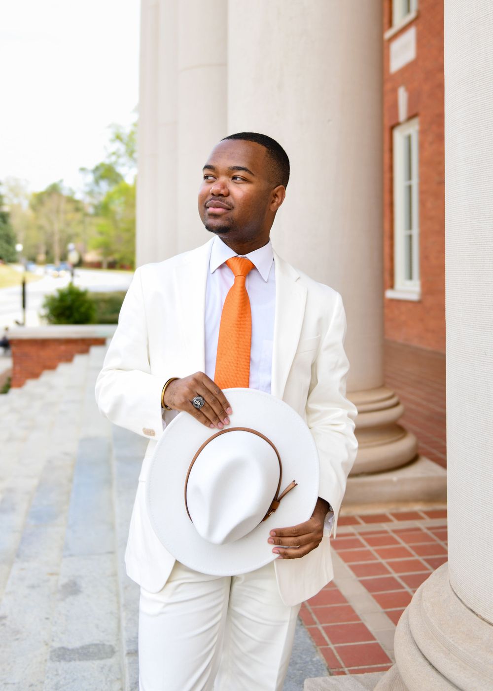 A person in a crisp white suit and orange tie stands confidently between classic columns holding a white hat.