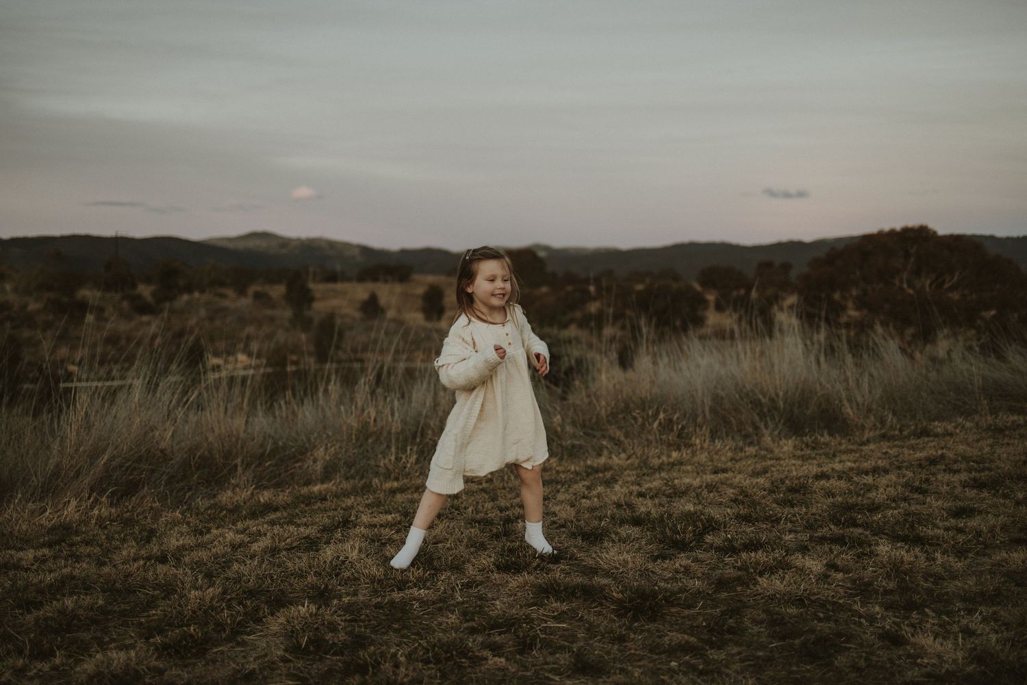 A girl in a flowing white dress twirls in a grassy field against a hilly Googong landscape at dusk.