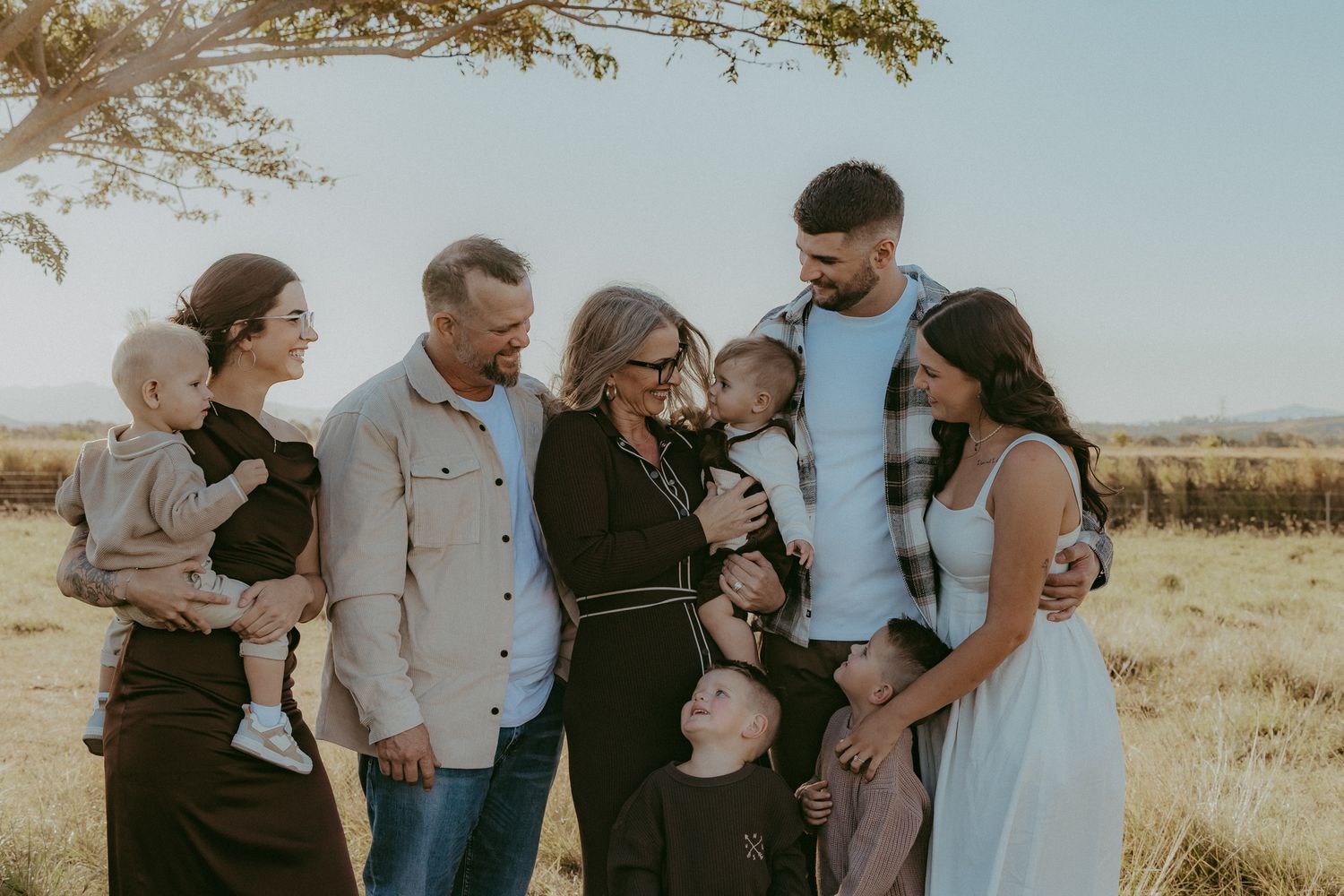Extended family sharing a warm embrace outdoors in a golden field with mountains visible in the background.