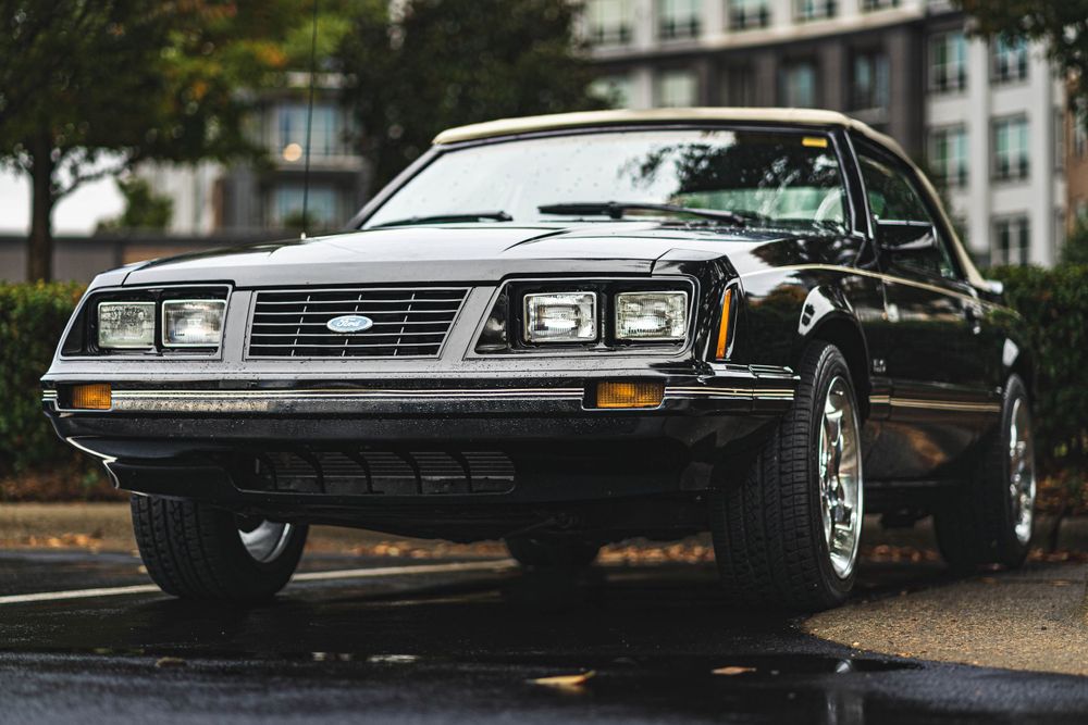 A classic black Mustang with retro quad headlights parked on a wet street.