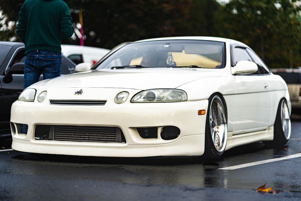 A lowered white Lexus SC300 with custom wheels parked on wet pavement.