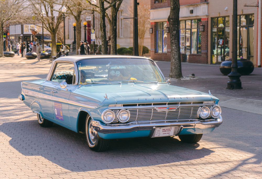 A classic light blue Chevrolet Impala parked on a tree-lined street in front of storefronts.