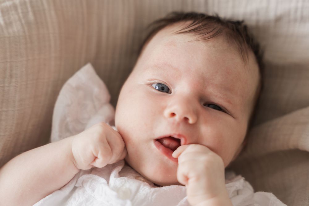 Baby lying on white bedding sucking thumb and looking at camera with bright eyes.