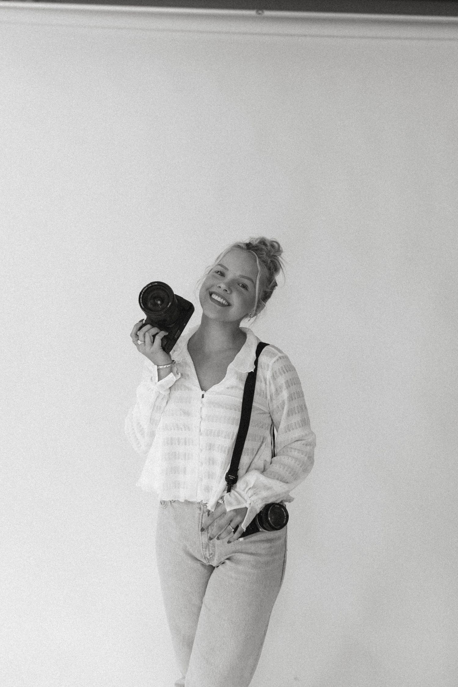 A person in a white button-up shirt and light jeans holds a camera while laughing against a white background.