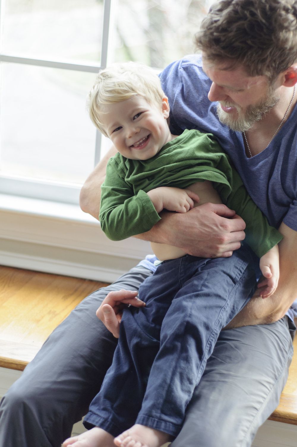Father playfully tickles his young son in their home window seat, capturing a joyful family moment.
