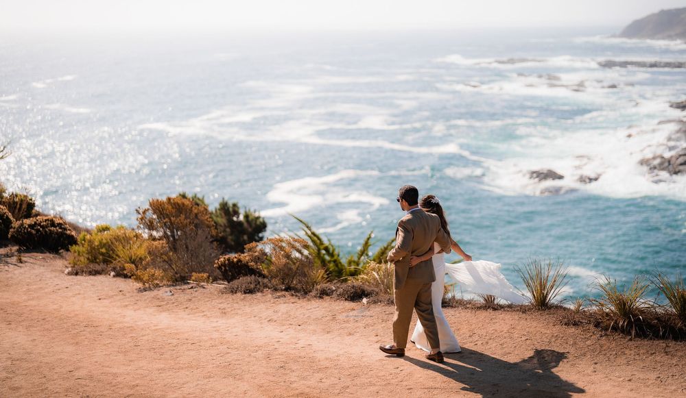 Couple embracing on a coastal cliff path overlooking turquoise ocean waves at Big Sur, California.