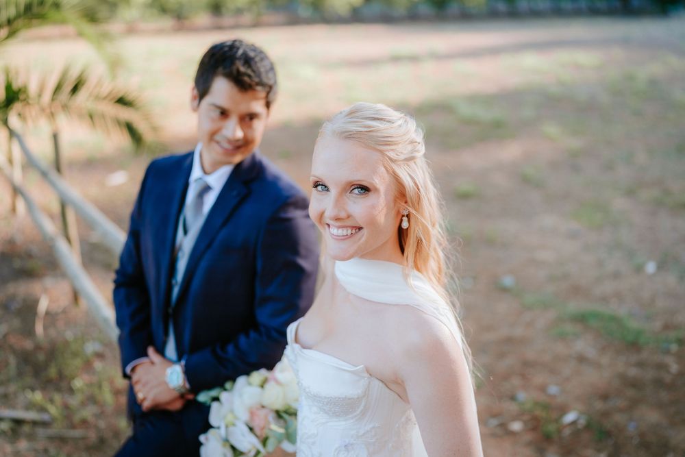 Smiling bride in white strapless gown holds bouquet outdoors, groom in navy suit stands behind her.