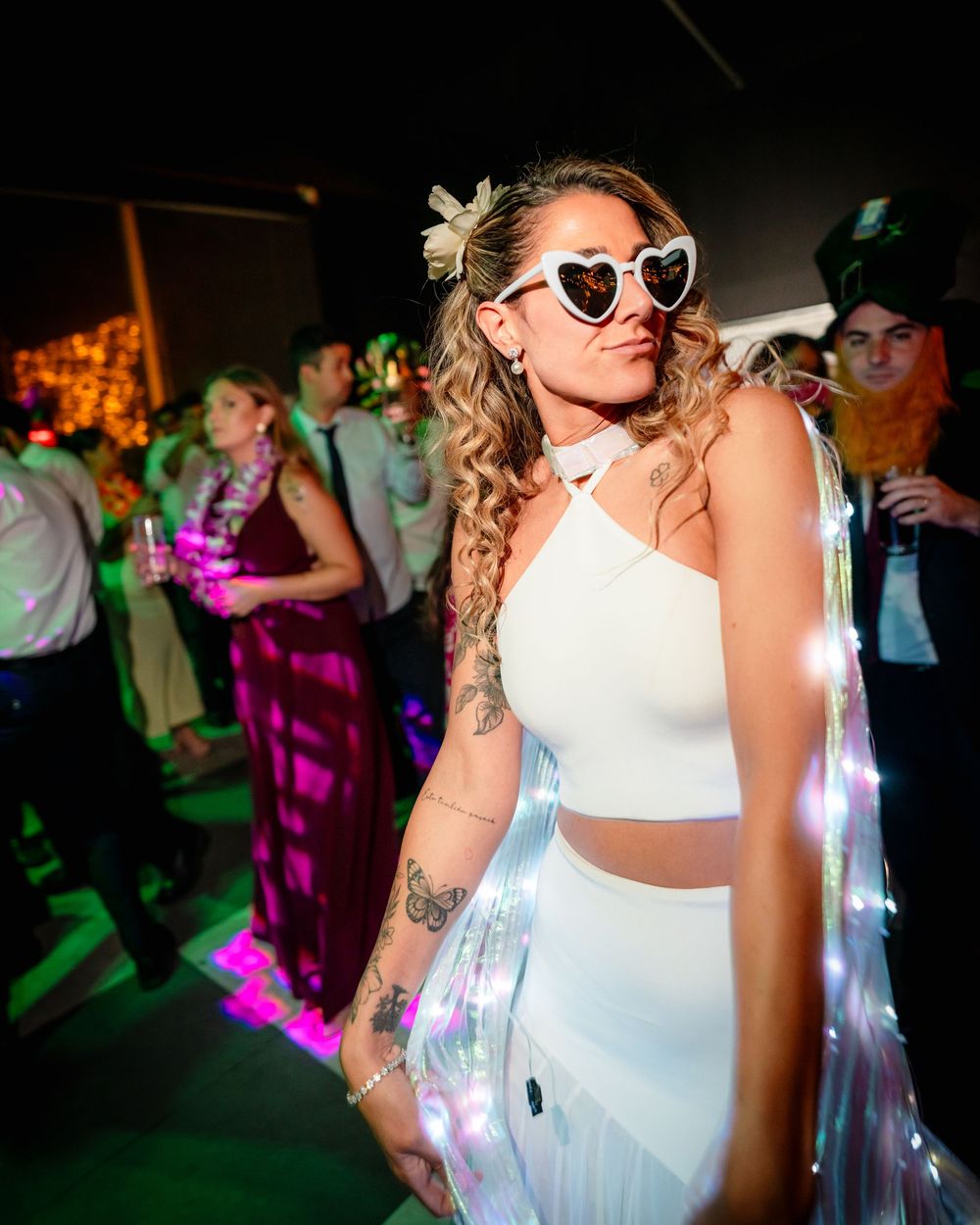 Tattooed bride in white crop top wearing heart sunglasses at vibrant wedding reception.