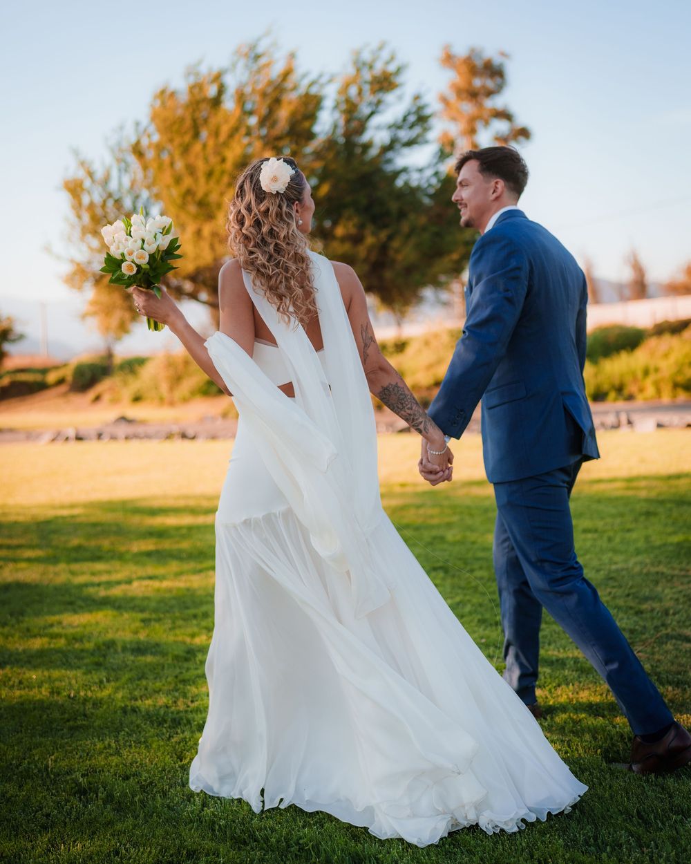 Bride in white gown holding bouquet walks hand in hand with groom in blue suit at golden hour.