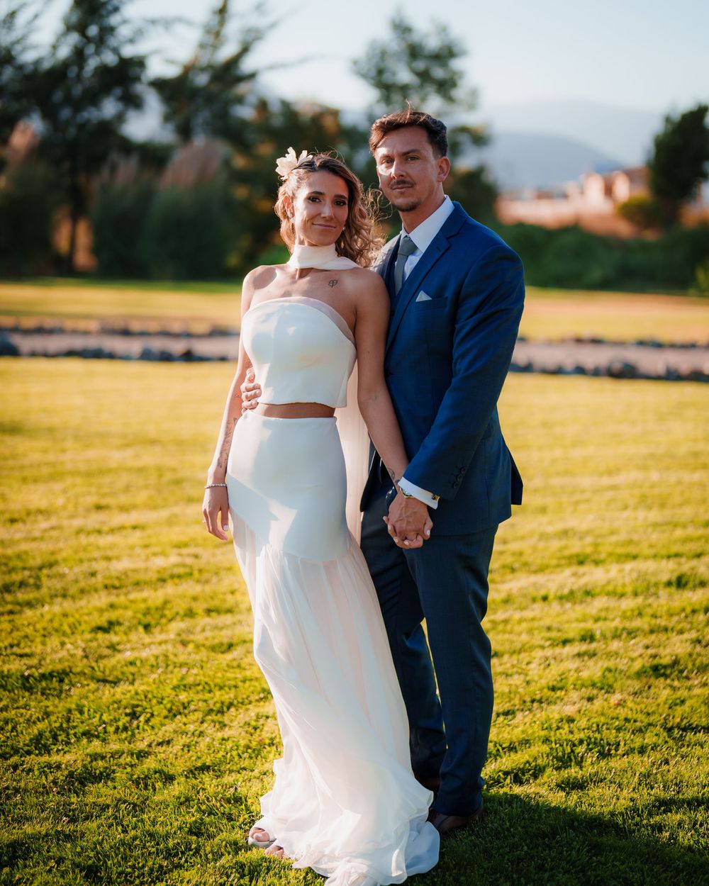 Couple in wedding attire pose together on green lawn with lavender fields and mountains in background.