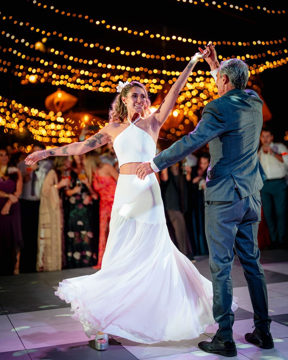 Bride in white two-piece gown dancing with groom under string lights at elegant wedding reception.
