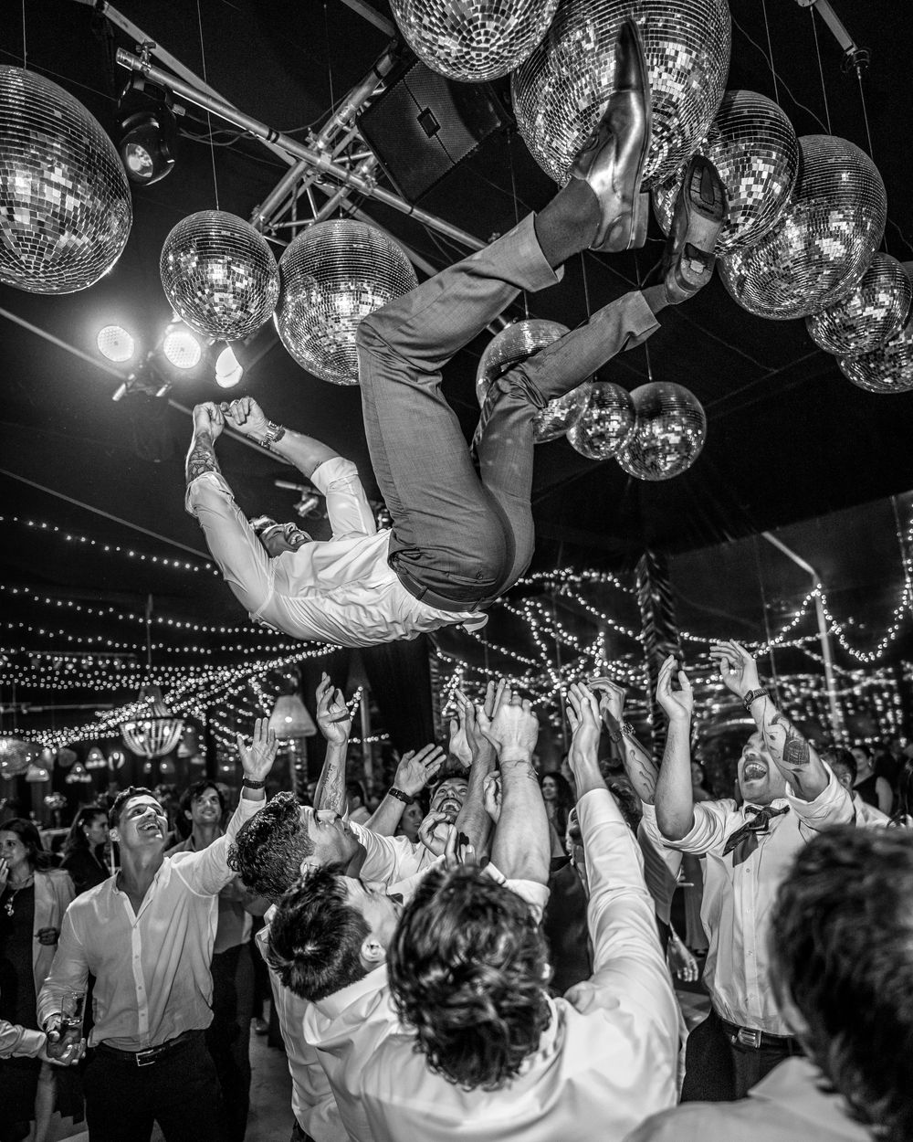 Groom being tossed in air by groomsmen under disco balls at lively wedding reception party.
