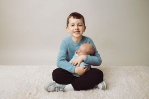 Child in blue sweater sitting cross-legged on light carpet while holding a sleeping newborn baby.