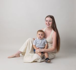 A parent and baby in matching neutral tones sit together on a light backdrop for a minimalist studio portrait.