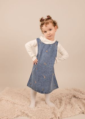 A child wearing a denim jumper dress with embroidered details and white long sleeve shirt poses on a beige background.