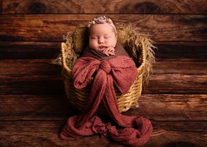 A newborn wrapped in a burgundy swaddle blanket lies in a rustic woven basket against a wooden backdrop.