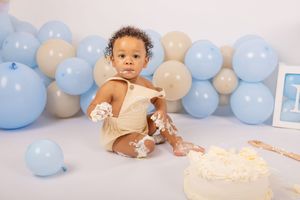 Baby in white outfit sits among blue and white balloons during cake smash photo session.