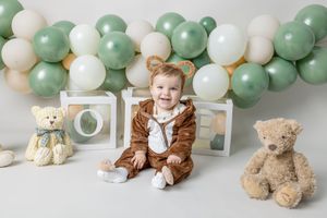 A series of baby photos and teddy bears arranged with sage green and white balloon decorations for a nursery or celebration.