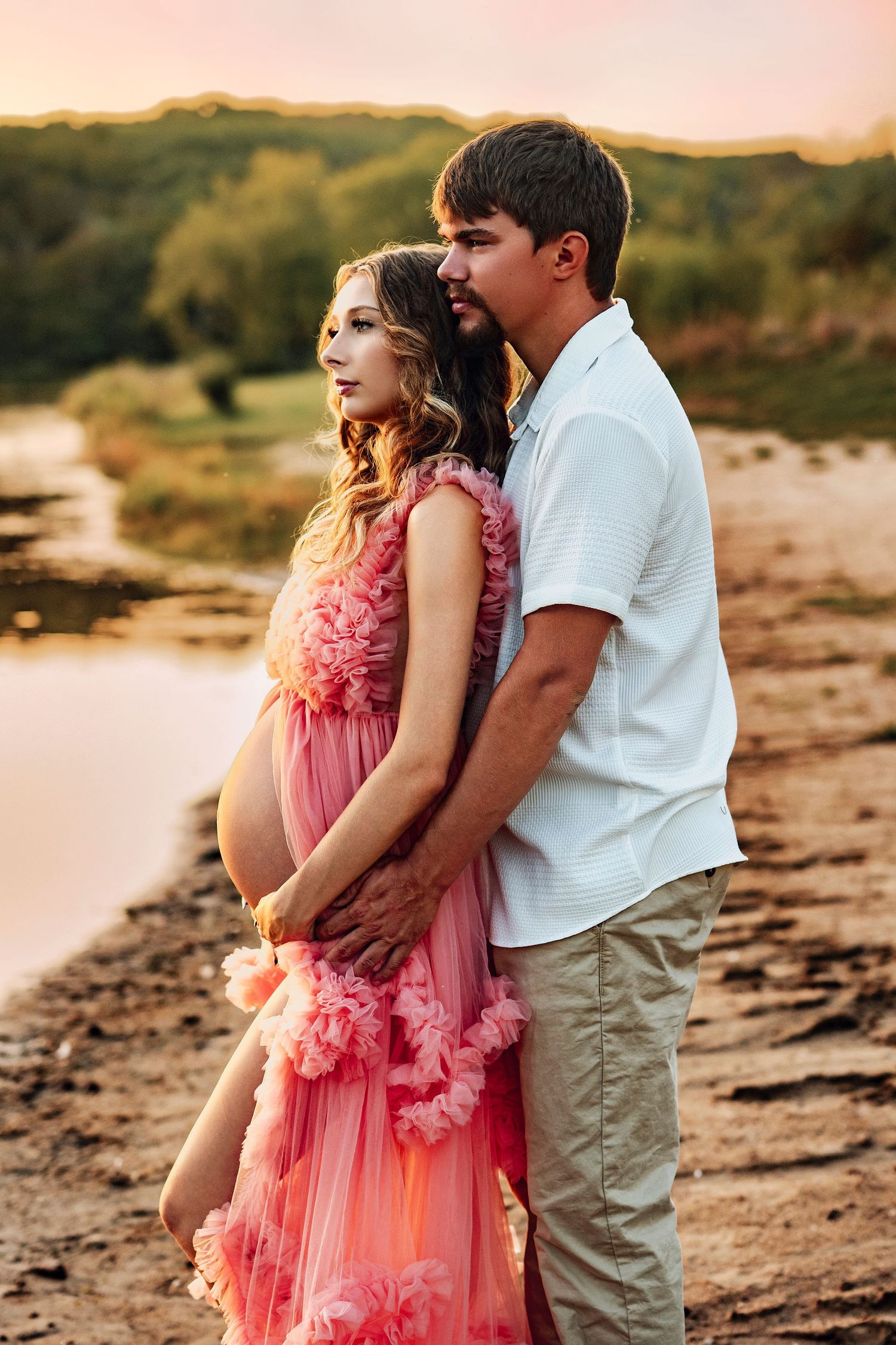 Pregnant woman in pink ruffled gown embraced by man in white shirt during golden hour by riverside. Nine Eagles Lake