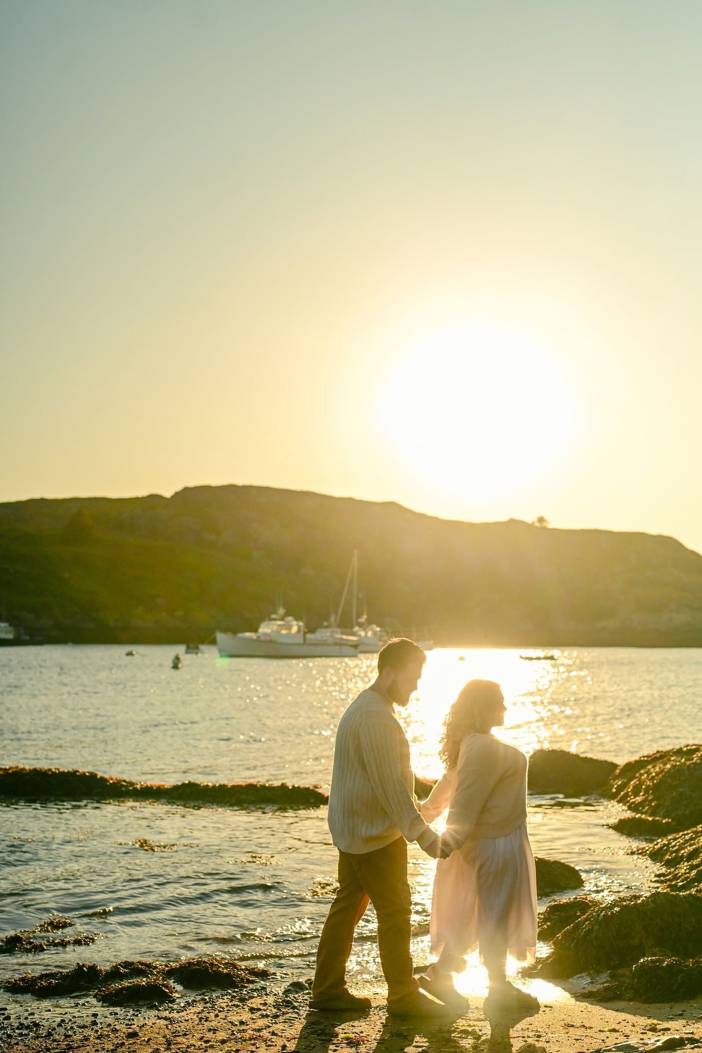Couple holding hands on rocky beach at golden sunset with sailboat and green hills in background.