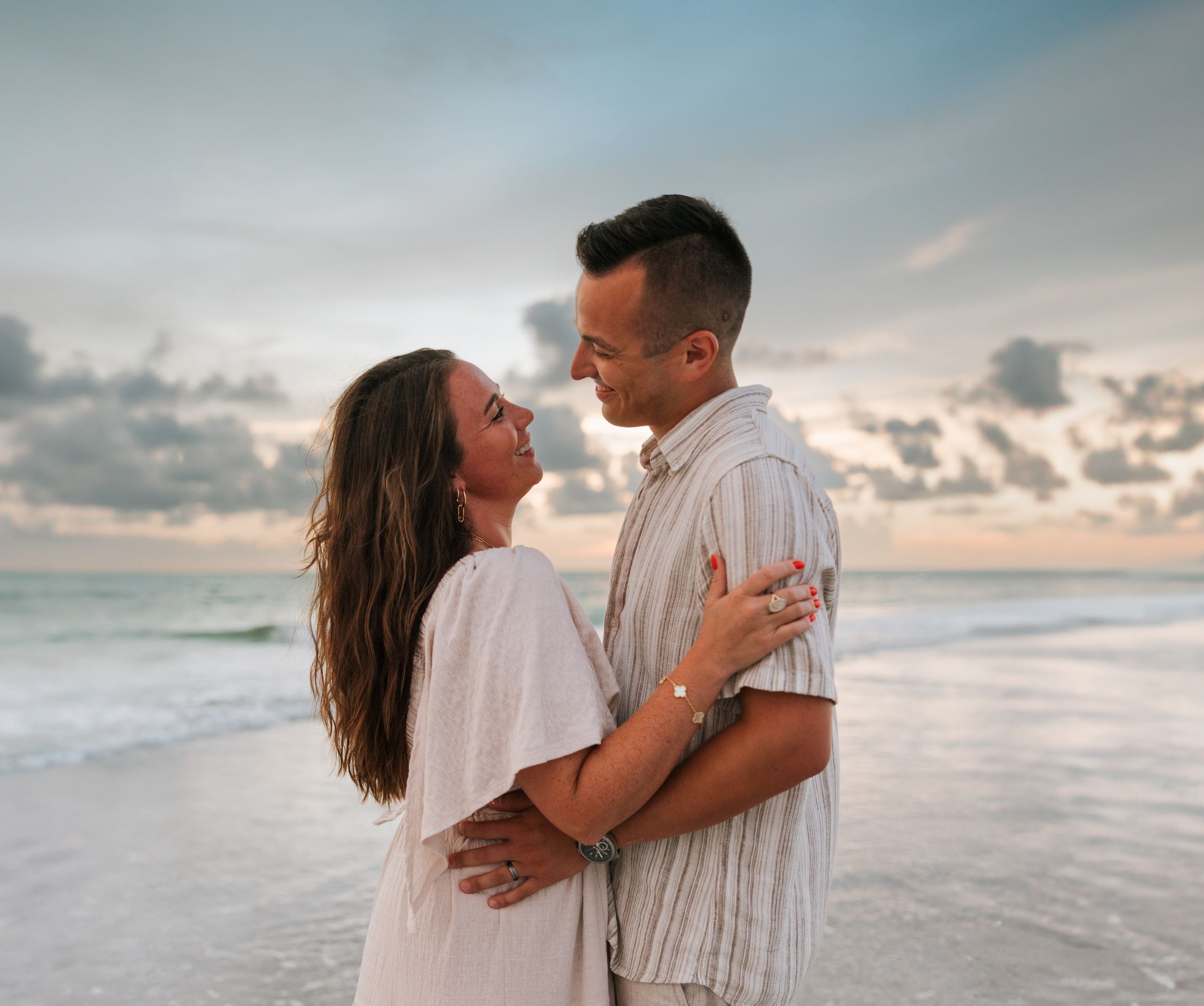 Romantic couple embracing on beach at sunset with ocean waves behind them.