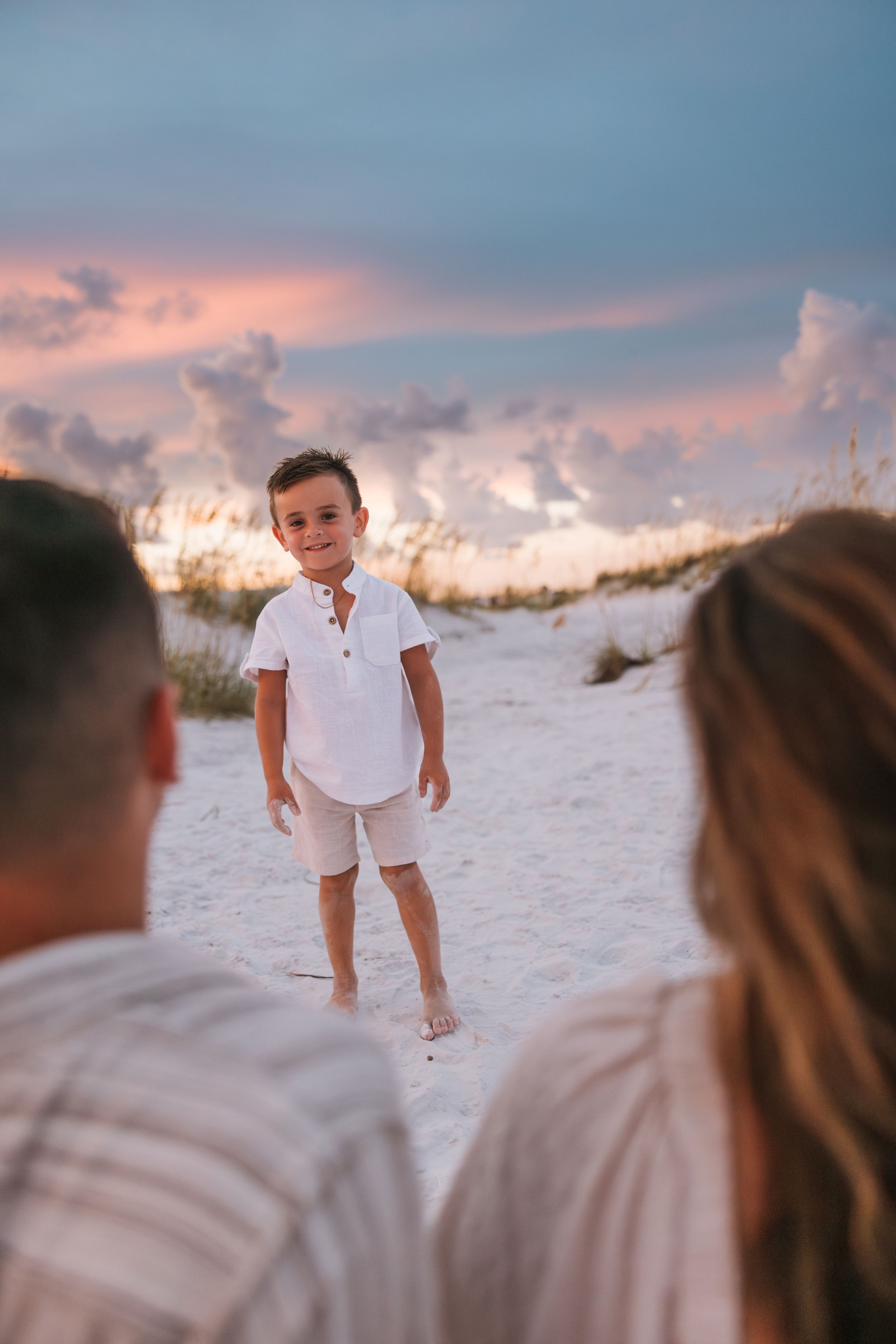 Child in white outfit stands on beach sand during sunset while parents look on.