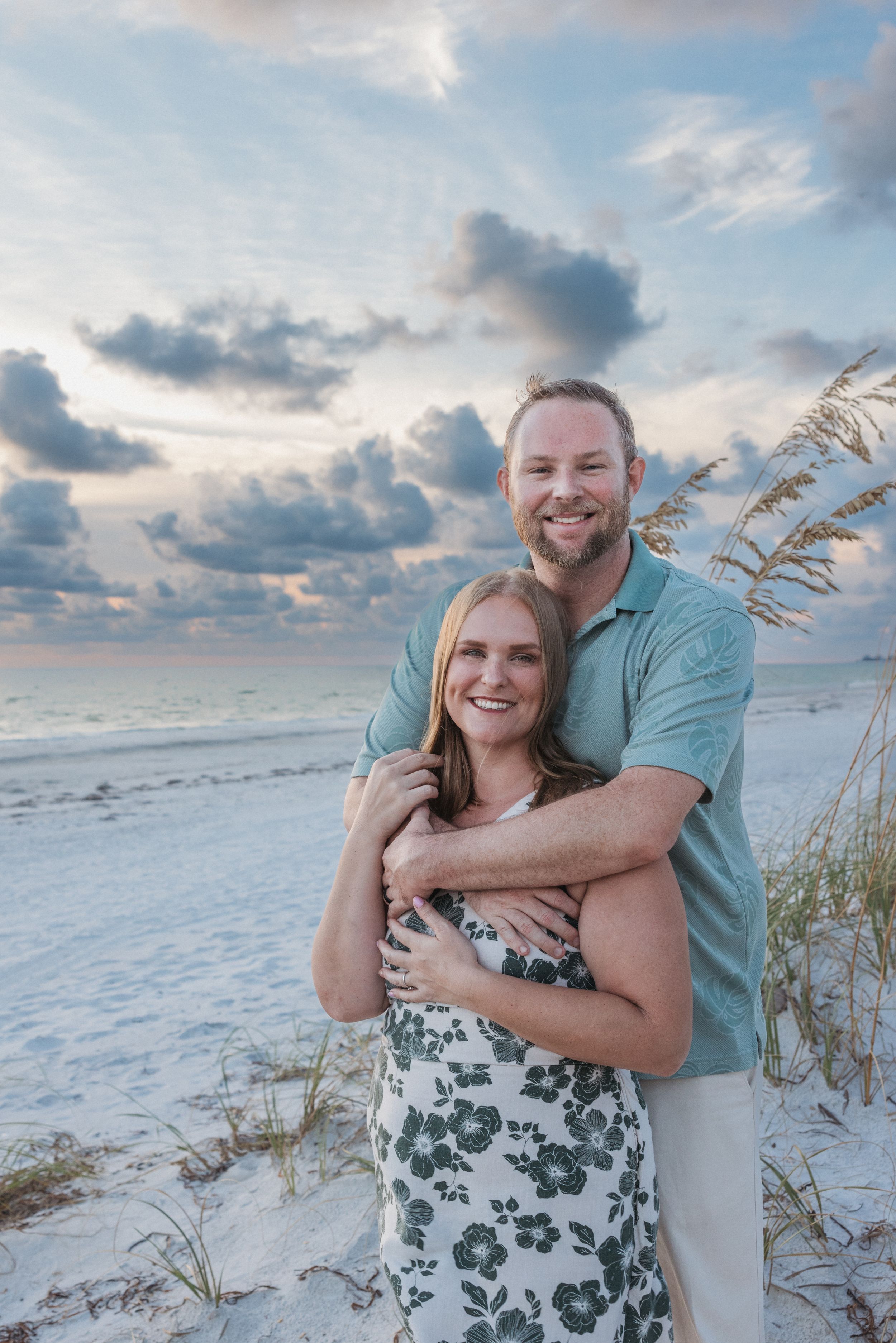 Couple embraces on beach dunes with sea oats and cloudy sunset sky.