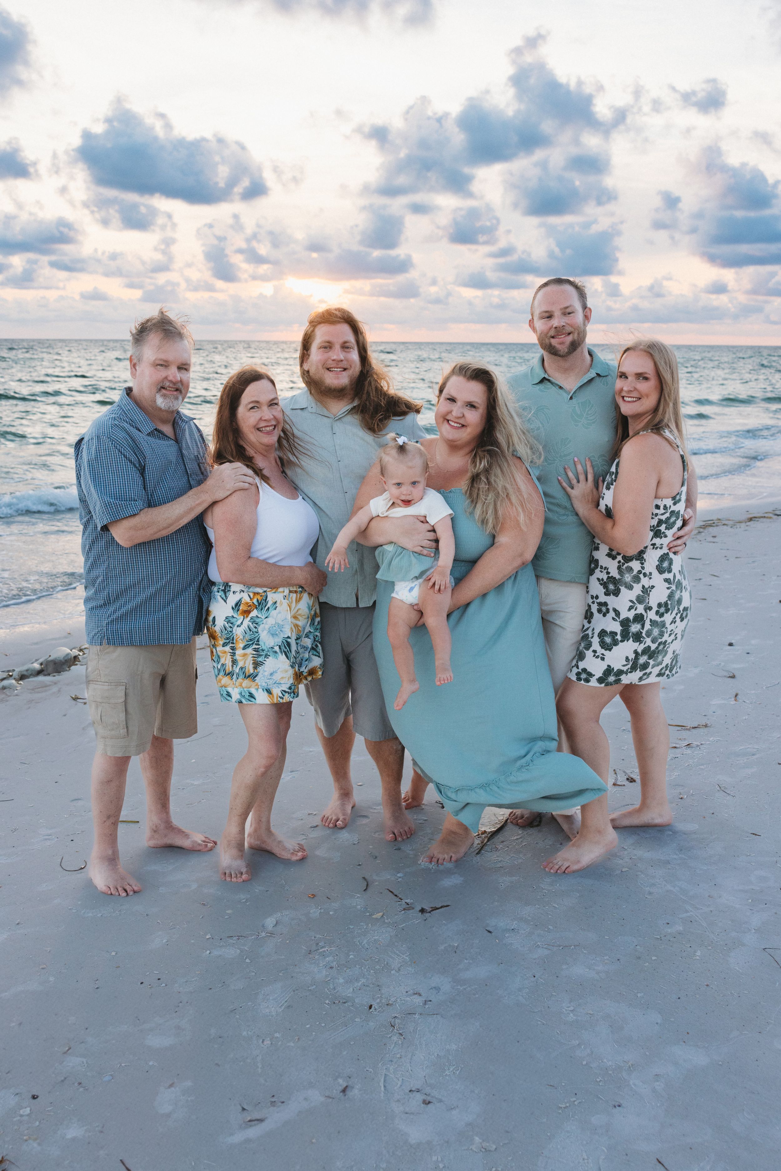 A cheerful family poses together on a beach at sunset, with cloudy skies and ocean waves in the background.
