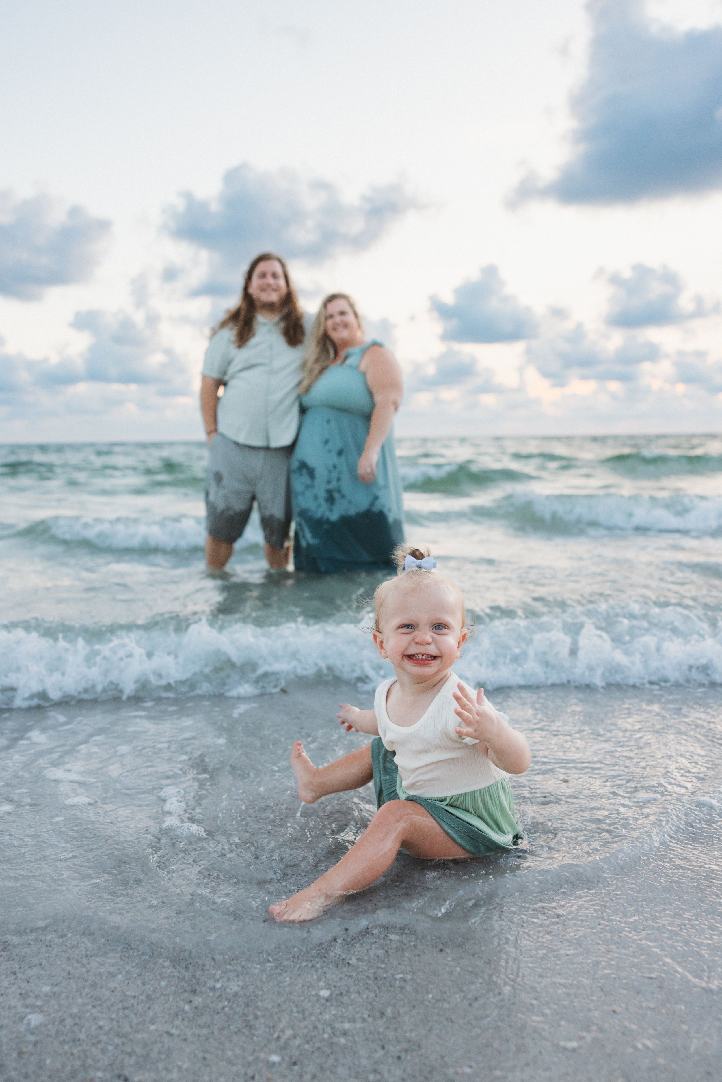 A toddler plays in the shallow ocean waves at sunset while people stand in the background.