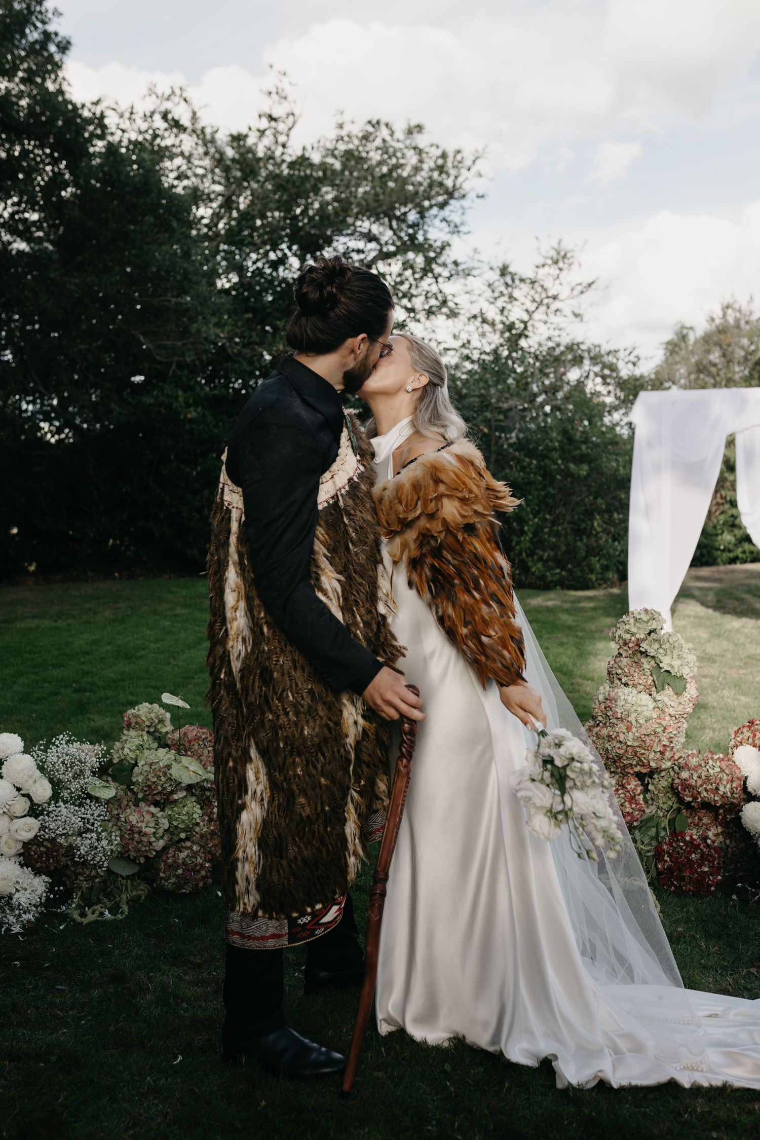 Bride and groom sharing first kiss outdoors with floral arch and garden backdrop.