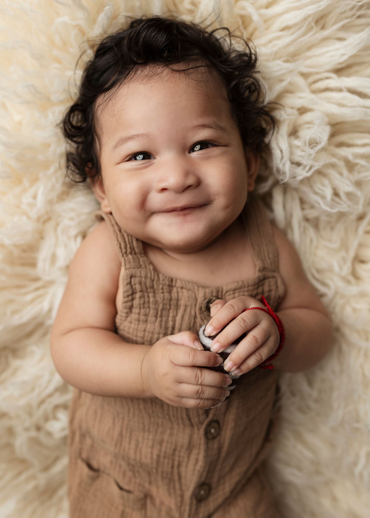 Baby in brown linen romper lying on cream fur backdrop.