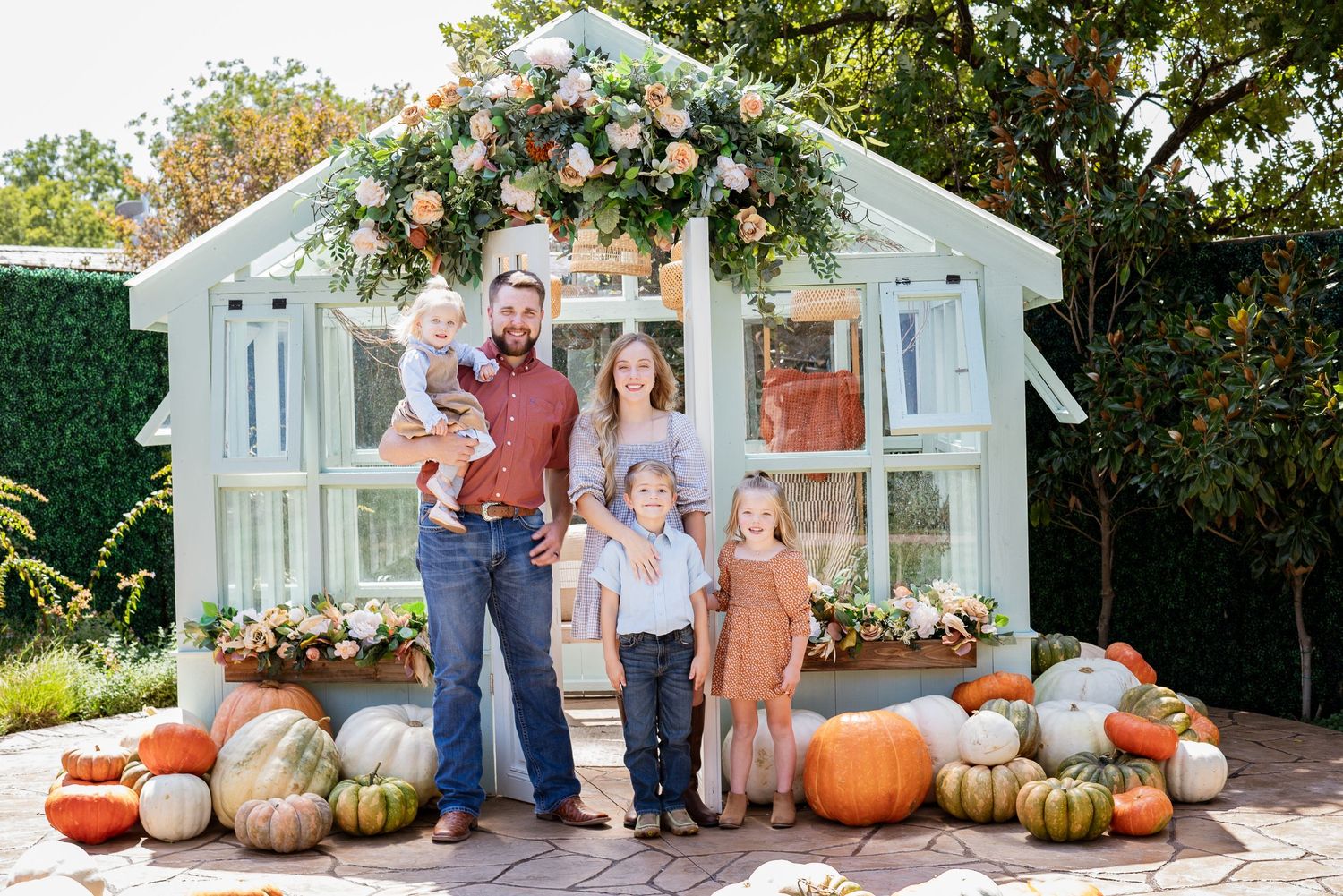 A family poses in front of a decorated greenhouse with fall pumpkins and floral arrangements.