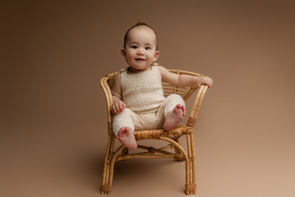 Baby wearing cream outfit sitting in rattan chair against brown background.