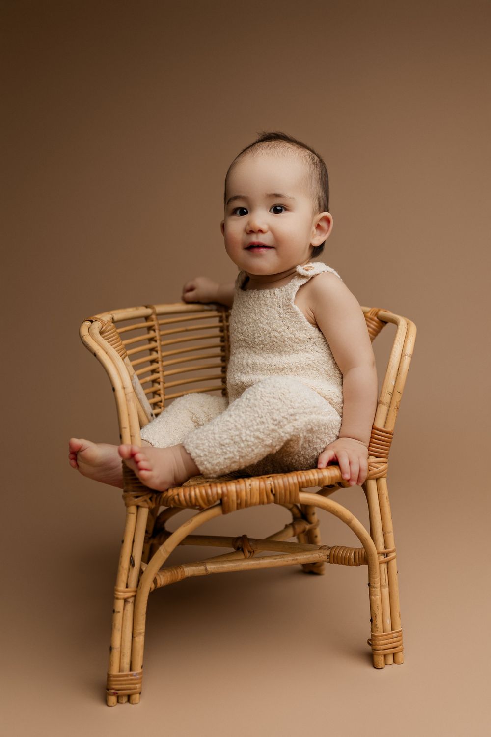 Baby wearing cream colored outfit sits in rattan chair against brown backdrop during professional photoshoot.