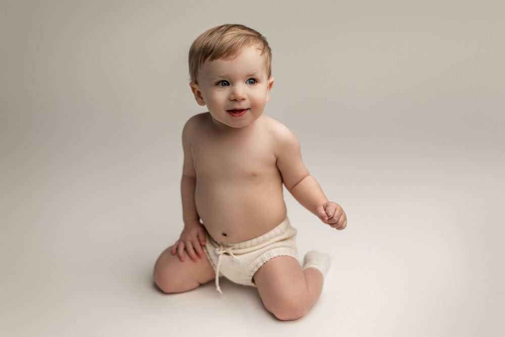 Baby in white diaper sits on seamless white background during professional portrait session.