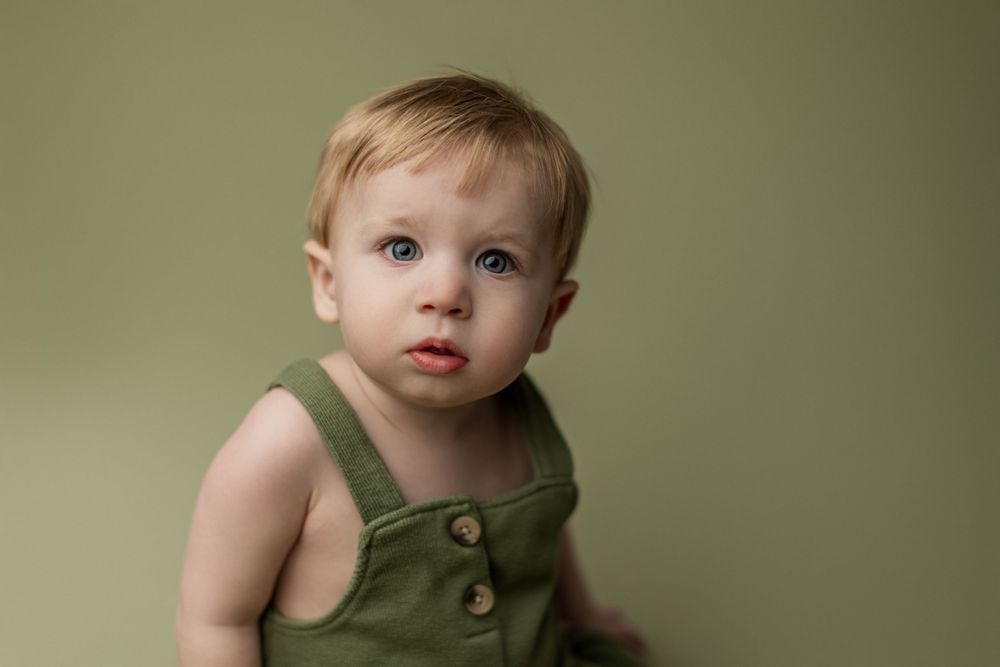 Baby in green overalls poses against olive background during professional portrait session.