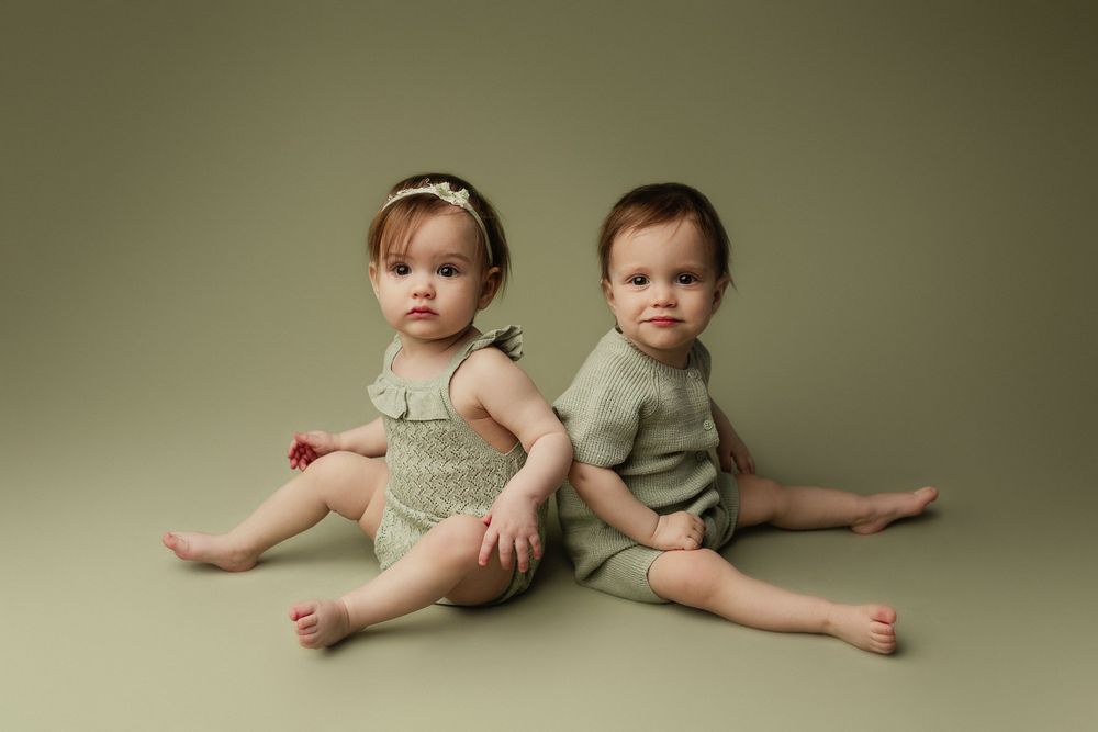 Two babies in matching sage outfits sitting together on beige background.