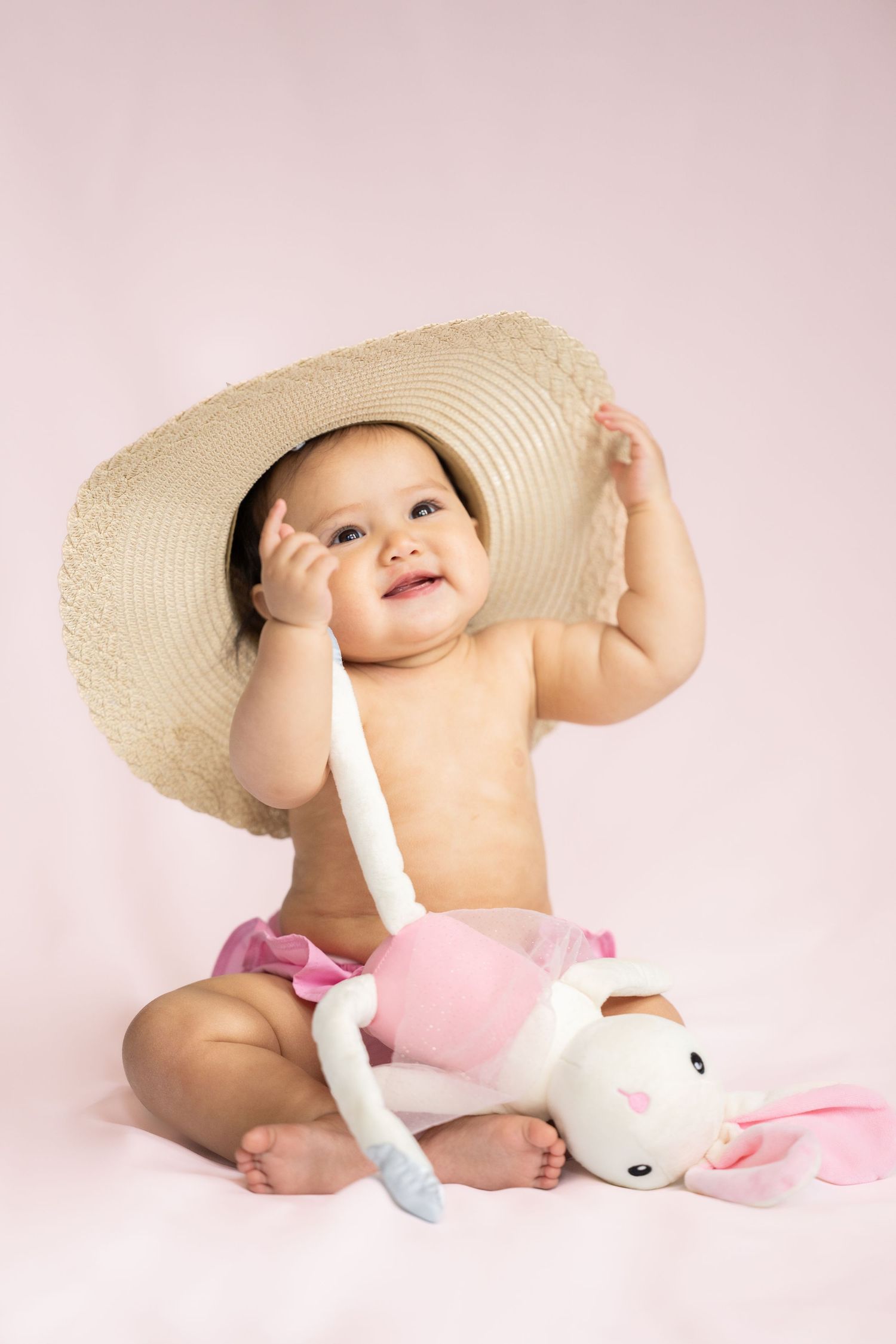 A smiling baby wearing a large sun hat sits with a stuffed bunny on pink background in studio.