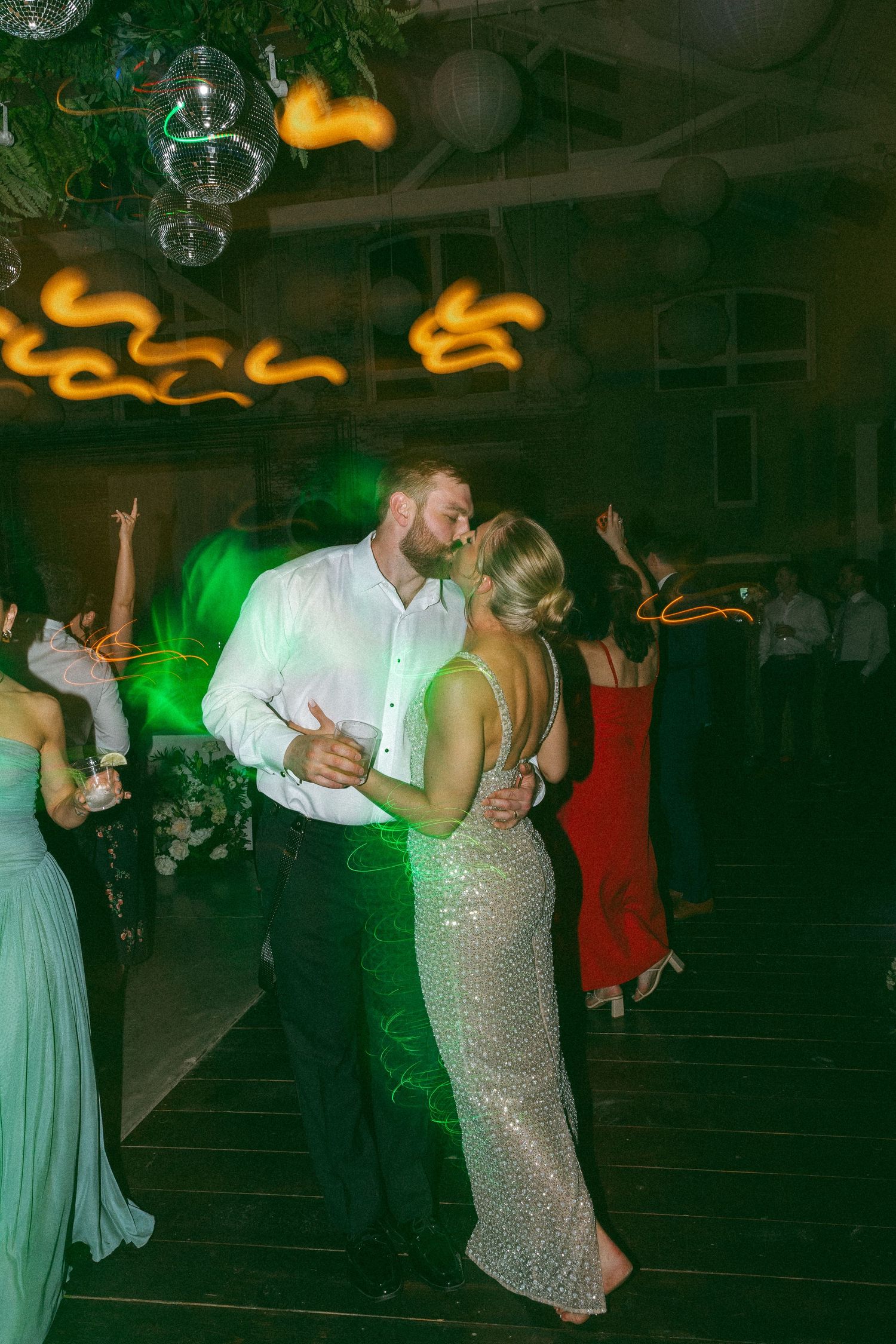 Couple shares a romantic first dance kiss at wedding reception with disco balls and colorful lights surrounding them.
